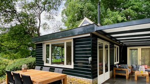 A modern house with a patio area featuring a wooden table and chairs, a built-in bench, and a glass door leading to a covered outdoor space. - Idyllic house with 360 view of nature and hottub (Dalfsen)