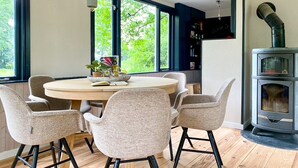 A dining area with a round table and six chairs, large windows with a view of greenery, and a wood-burning stove. - Idyllic house with 360 view of nature and hottub (Dalfsen)