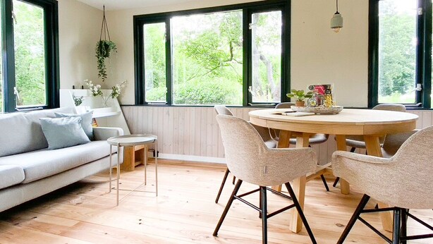 A modern living room with a round wooden table, two chairs, a sofa, and large windows with a view of greenery. - Idyllic house with 360 view of nature and hottub (Dalfsen)