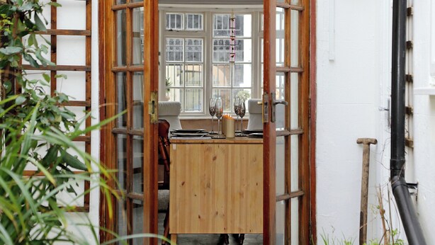 Une porte en bois avec des panneaux de verre qui donne sur une pièce où une table est dressée pour le repas, avec un mur blanc et une lanterne suspendue.