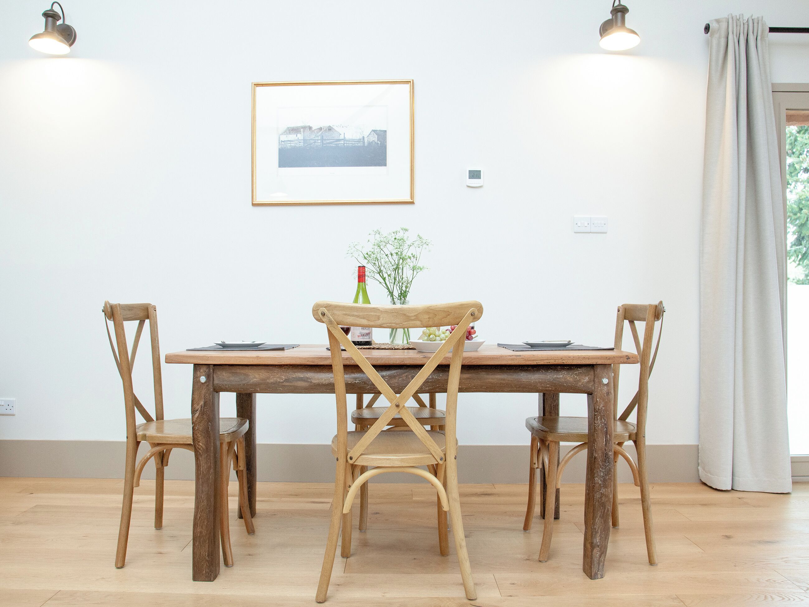 A dining area with a wooden table and four chairs, a framed picture on the wall, and a window with curtains.