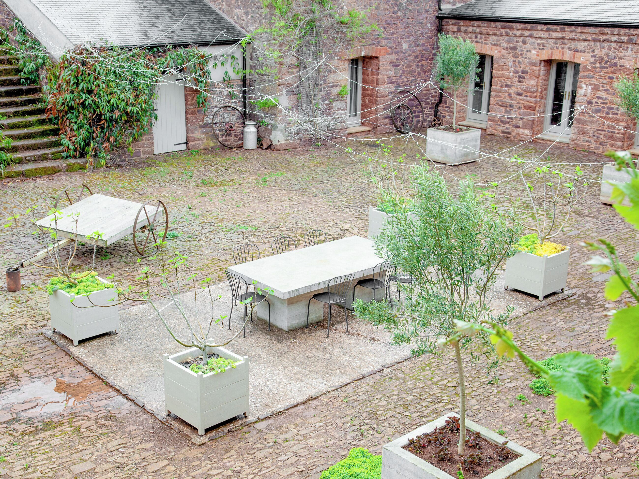 Un patio con suelo de piedra, una mesa de madera con sillas y varios maceteros con plantas.