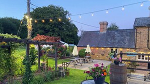 Une terrasse de jardin avec des tables et des chaises en bois, des guirlandes lumineuses et un bâtiment rustique doté d’une cheminée en briques.