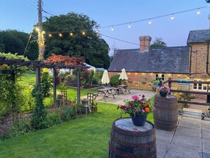 A garden patio with wooden tables and chairs, string lights, and a rustic building with a brick chimney. - The Old Kitchen -UK32062 (Burwarton, near Bridgnorth)