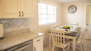 Una cocina con armarios blancos, encimera de granito, una mesa de comedor con sillas blancas y un reloj de pared.