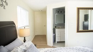 Un dormitorio con cama, lámpara de noche, ventana, espejo y un baño visible a través de una puerta abierta.