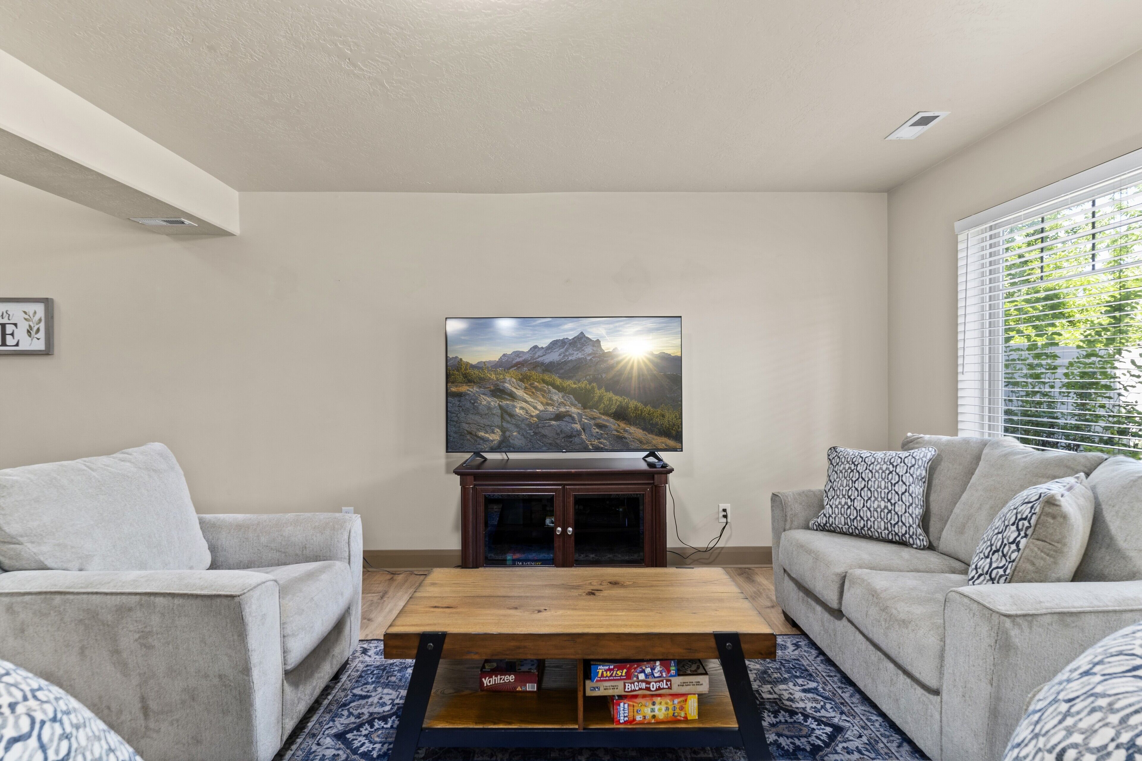 Una sala de estar con un televisor de pantalla plana montado en la pared, dos sofás grises, una mesa de centro de madera y una ventana con persianas.