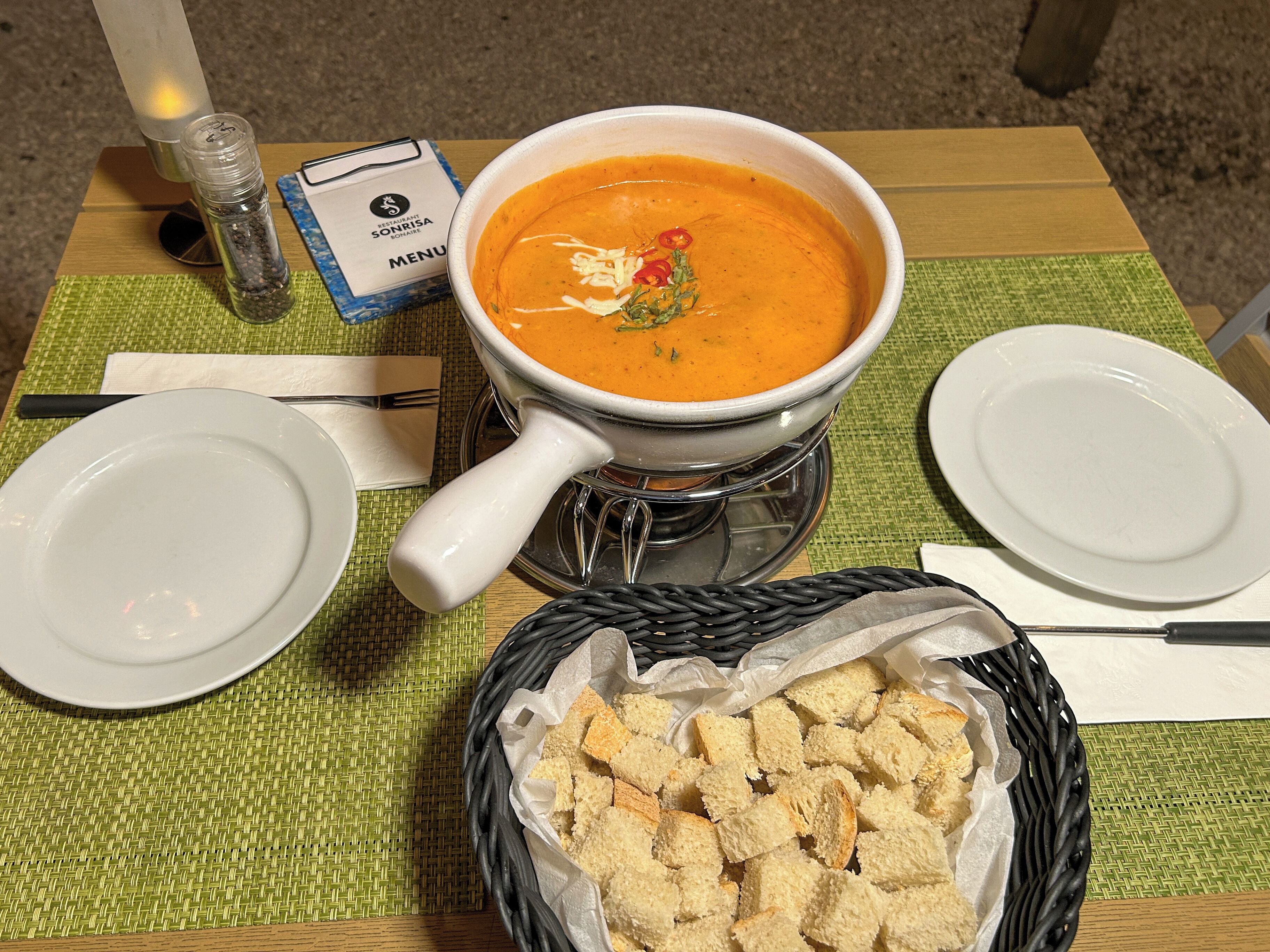Une table avec un bol de soupe, un menu, un salier, un panier de pain et deux assiettes.