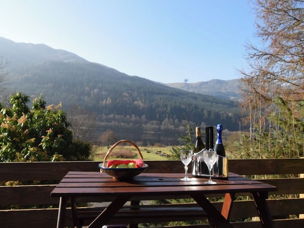 A wooden table with a basket of fruit, two wine glasses, and two bottles, set against a mountainous backdrop.