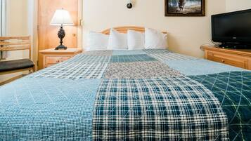 A neatly made bed with a blue and white checkered bedspread and a quilt. There is a wooden nightstand with a lamp and a television on it. A wooden chair is visible to the left of the bed.