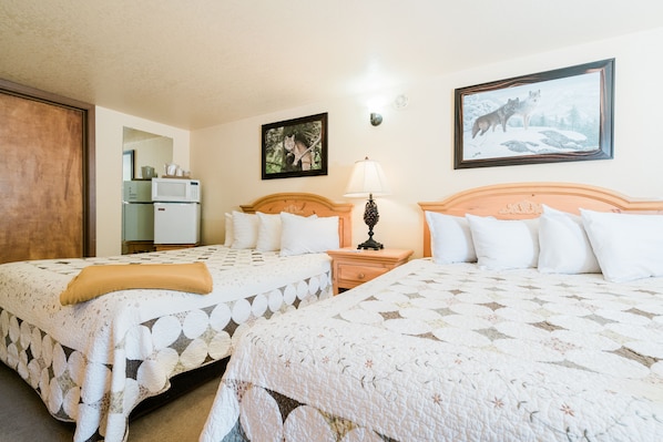 A hotel room with two beds, a wooden headboard, a nightstand with a lamp, a refrigerator, a microwave, a framed picture on the wall, and a door.