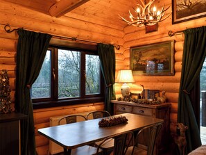 A wooden cabin interior with a dining table, chairs, a chandelier, a window with curtains, a lamp, and a framed picture on the wall.