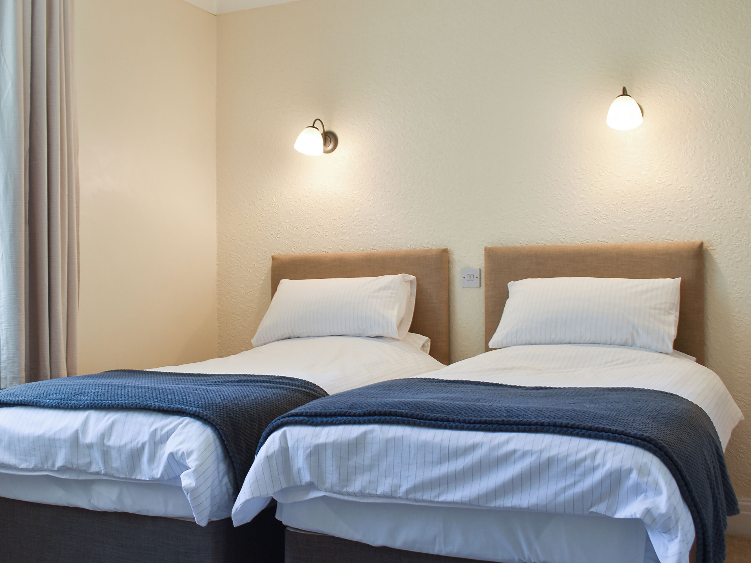 Une chambre d’hôtel avec deux lits, chacun doté d’une tête de lit, de draps blancs et d’une couverture bleue. Des lampes murales sont fixées au-dessus de chaque lit.
