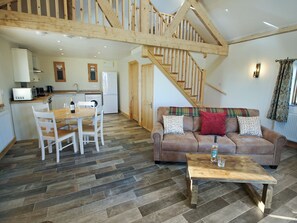 A spacious living area with a wooden staircase, a dining table with chairs, a sofa with patterned cushions, and a kitchen in the background.