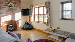 A living room with a brick fireplace, a flat-screen TV mounted on the wall, a window with curtains, and a wooden floor. - Tawny Cottage - UKC2580 (Yoxall, near Lichfield)