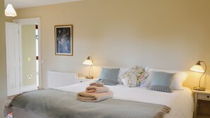 Een netjes opgemaakt bed met een deken, kussens en een opgevouwen handdoek. Twee nachtlampjes, een ingelijst schilderij aan de muur, en een witte deur die naar een andere kamer leidt.