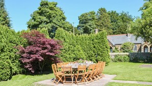 A round wooden table with chairs set outdoors, surrounded by lush greenery and a stone wall.