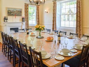 Comedor con una larga mesa dispuesta para una comida, sillas, chimenea y grandes ventanales.