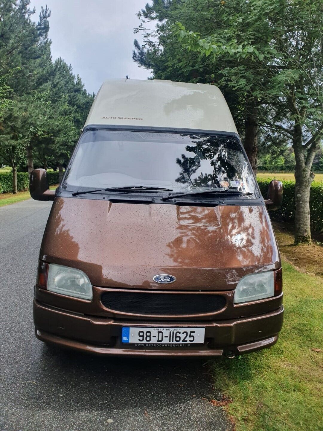 A brown Ford van with a camper shell parked on a road surrounded by trees.