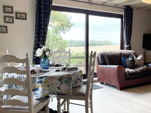 A dining area with a table set for two, a sofa, and a large window with a view of the outdoors. - 2 bedroom accommodation in Southrepps (Southrepps)