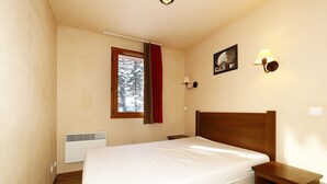 A bedroom with a wooden bed, a nightstand, a wall-mounted lamp, a window with a view of trees, and a radiator. - Appartement T2 Albane Vars (vars)