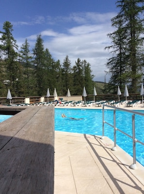 Piscina com deck de madeira em primeiro plano, rodeada por espreguiçadeiras e guarda-sóis.
