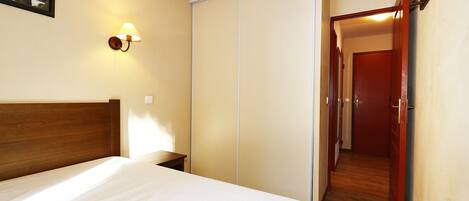A hotel room with a bed, a wardrobe, a nightstand, a wall-mounted lamp, and a door leading to a hallway.