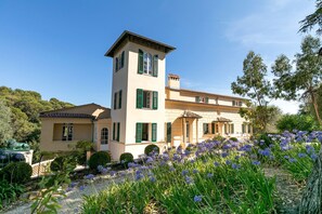 A two-story building with a tower, green shutters, and a garden with purple flowers.