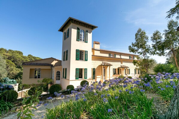 A two-story building with a tower, green shutters, and a garden with purple flowers.