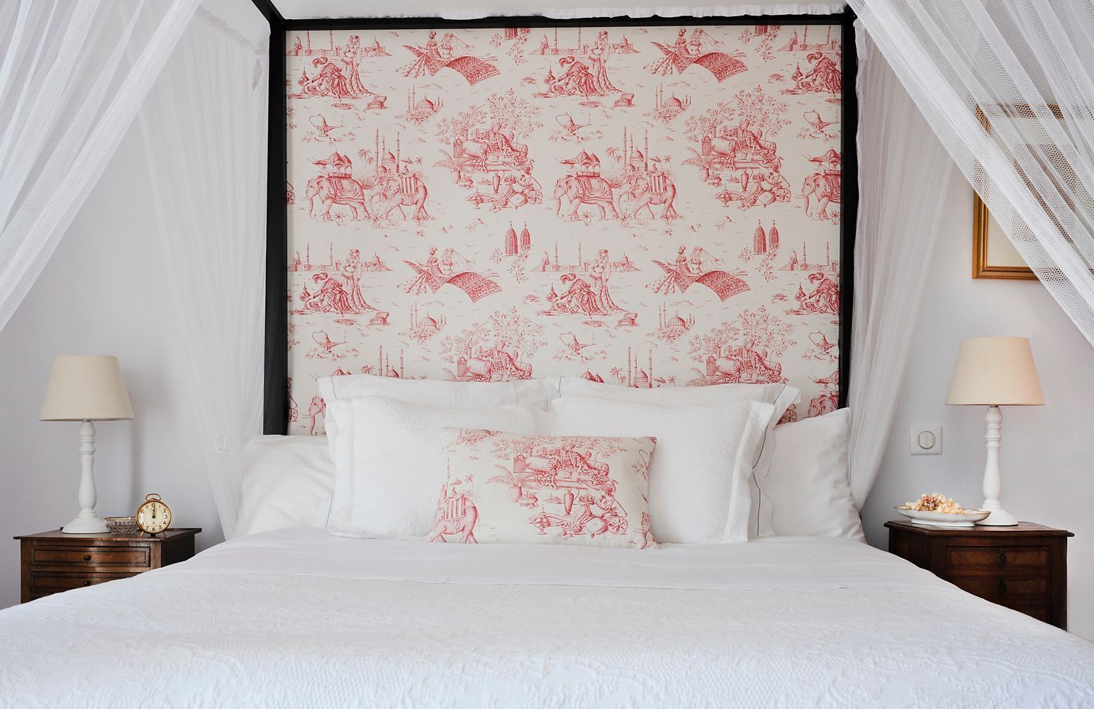 Uma cama com dossel e cabeceira estampada, duas mesas de cabeceira com abajures, e um relógio em uma delas.