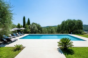A rectangular swimming pool with lounge chairs and umbrellas, surrounded by greenery and trees.
