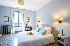 A hotel room with a bed, bedside lamps, a radiator, a window with curtains, and a picture on the wall.