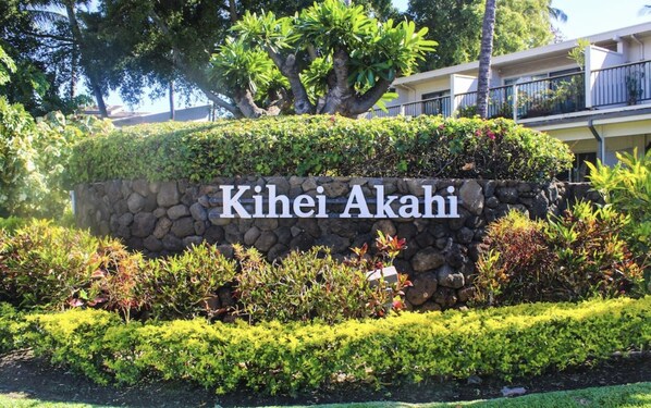 A stone wall with the text "Kihei Akahi" surrounded by lush greenery and a building in the background.