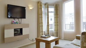 Un salon moderne avec un téléviseur à écran plat fixé au mur, un meuble de rangement en bois en dessous, une petite table avec un livre et une télécommande, un canapé et de grandes fenêtres avec des rideaux.