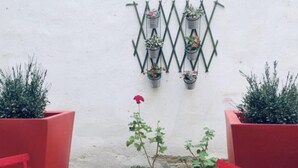 Un petit espace extérieur aménagé avec une table et des chaises rouges, entouré de plantes en pot et agrémenté d’un élément décoratif en métal fixé au mur.