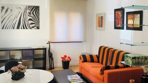Un salon moderne comprenant un canapé, une table basse et un coin repas avec des chaises et une table. Les murs sont ornés d’œuvres d’art et une grande fenêtre laisse entrer la lumière naturelle.