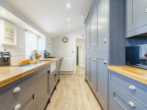 A modern kitchen with wooden countertops, grey cabinets, a clock on the wall, and a view into another room.