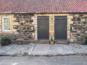 A stone building with two dark wooden doors, a window with a grid pattern, and a small planter with greenery. - Haven Cottage - E4342 (Holy Island)
