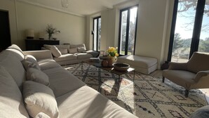 A spacious living room with a large beige sofa, a coffee table, and a patterned rug. There are two large windows providing natural light and a view of the outdoors. - Bastide sleeps 9, large secure swimming pool, not overlooked in Aix en Provence (Aix-en-Provence)