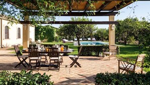 An outdoor patio with a pool, surrounded by greenery and furnished with wooden chairs and tables.