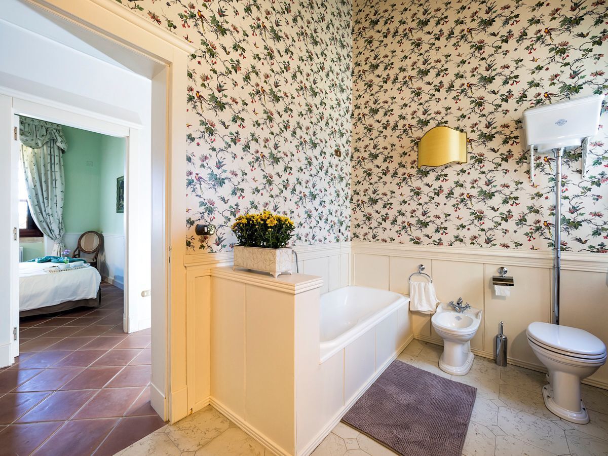 Une salle de bain avec une baignoire, des toilettes et un bidet. Les murs sont recouverts d’un papier peint à motifs floraux et il y a une lampe dorée.