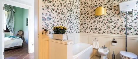 Une salle de bain avec une baignoire, des toilettes et un bidet. Les murs sont recouverts d’un papier peint à motifs floraux et il y a une lampe dorée.