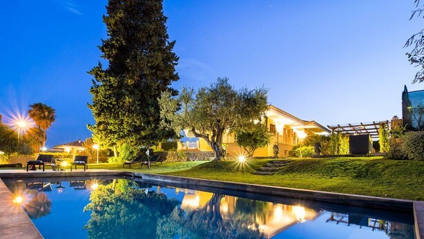 A swimming pool with a clear reflection, surrounded by well-lit outdoor seating and a large tree.