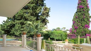 A balcony with a white cushioned seating area, a small table with a teapot and cups, and a view of a garden with cacti and trees.