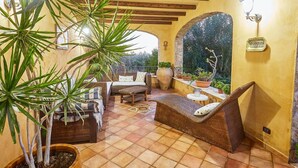 Une terrasse ensoleillée, meublée de meubles en osier, agrémentée de plantes en pot et surmontée d’une arche en pierre.