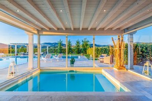 A modern indoor pool area with clear glass walls, wooden ceiling beams, and a view of a landscaped outdoor area with lounge chairs and a pool. - Villa Ferdinando - Five Bedroom Villa, Sleeps 10 (Pieve a Nievole)