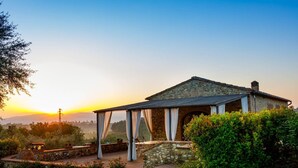 A stone building with a covered patio, surrounded by greenery and a clear sky.
