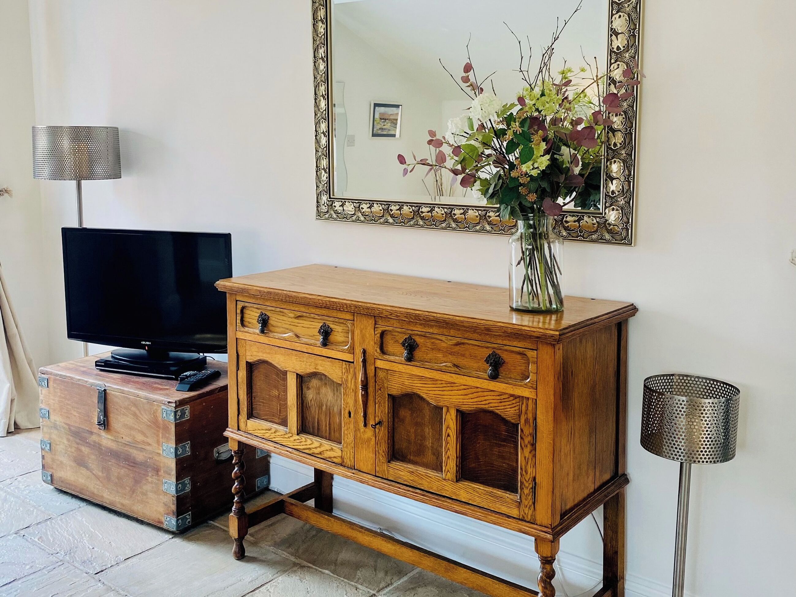 Ein Holzkabinent mit Schubladen, ein Fernseher auf einem Sockel, eine Vase mit Blumen, ein Spiegel mit aufwendigem Rahmen und eine Lochlampe.