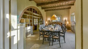 Un espace repas avec une longue table dressée pour un repas, entourée de chaises. On y trouve une bibliothèque remplie de livres et d’objets décoratifs, ainsi qu’une grande fenêtre qui laisse entrer la lumière naturelle.