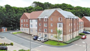 A multi-story residential building with a brick facade, multiple windows, and a parking area with several cars.